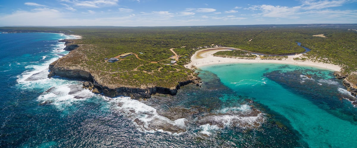An aerial view of a coastal landscape with a beach, clear turquoise waters, and a dense forested area.