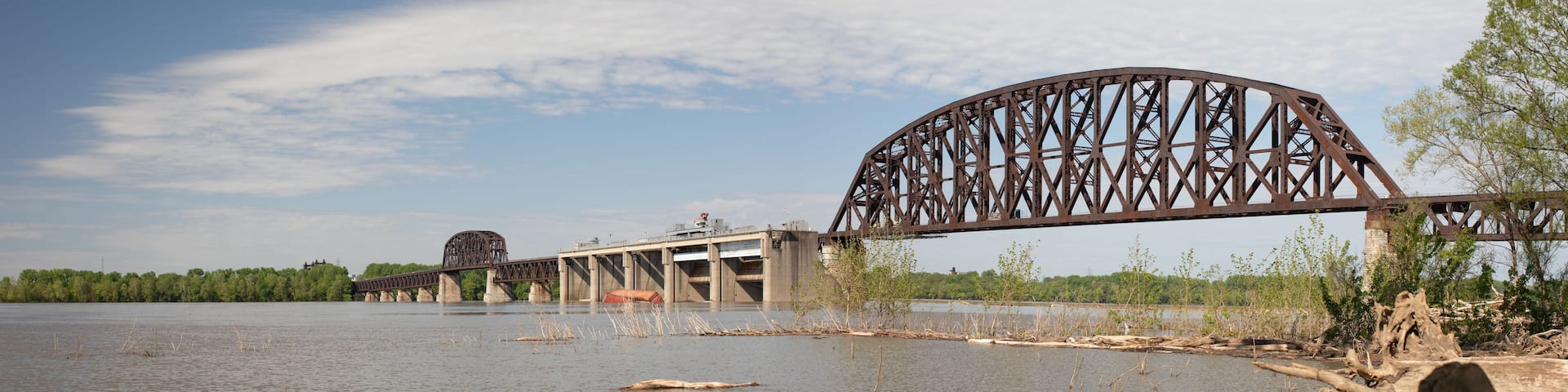 A large steel bridge over a river with trees and a cloudy sky in the background.
