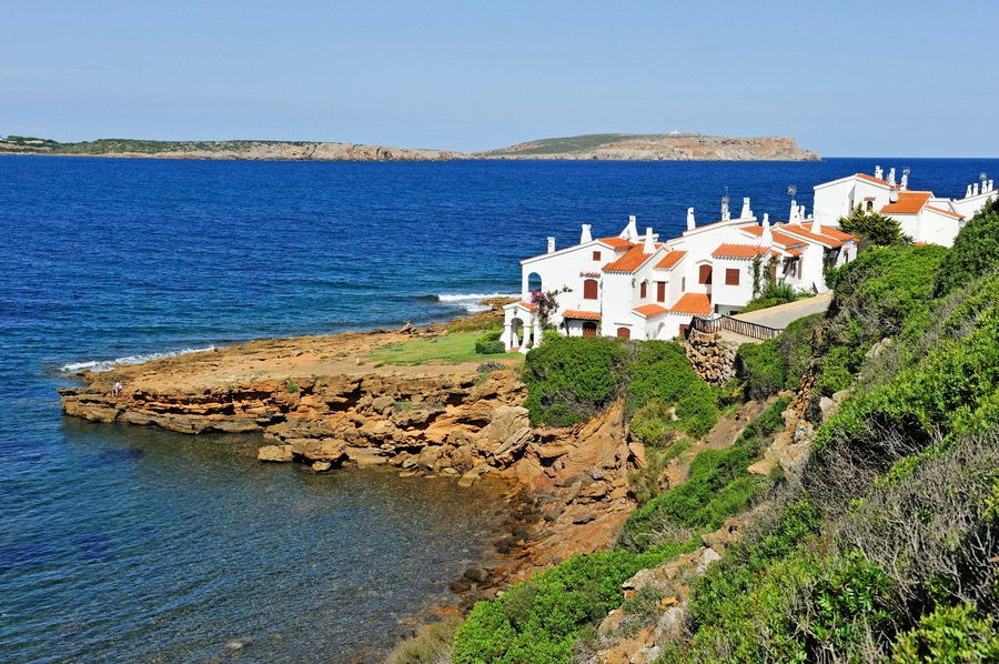 A coastal village with white buildings and red roofs, rocky cliffs, and the sea.
