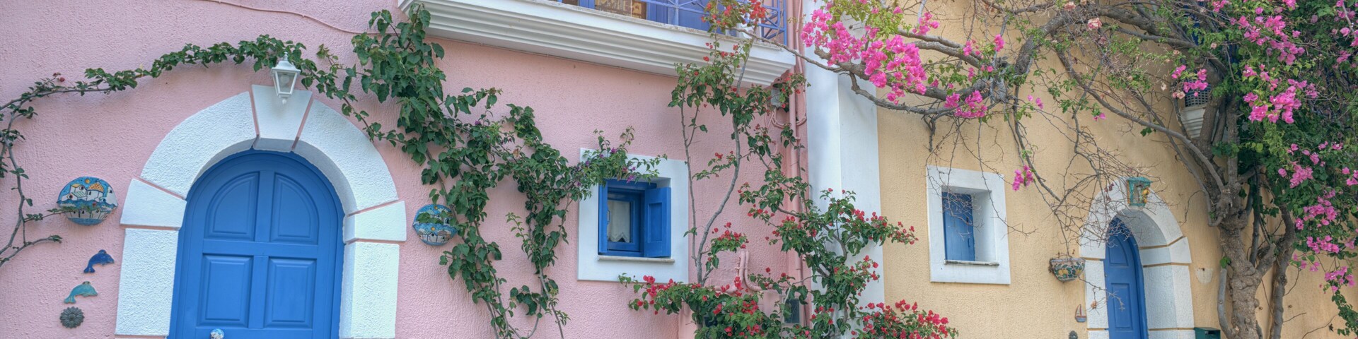 A pink and yellow building with blue doors and windows, surrounded by flowering plants and vines.