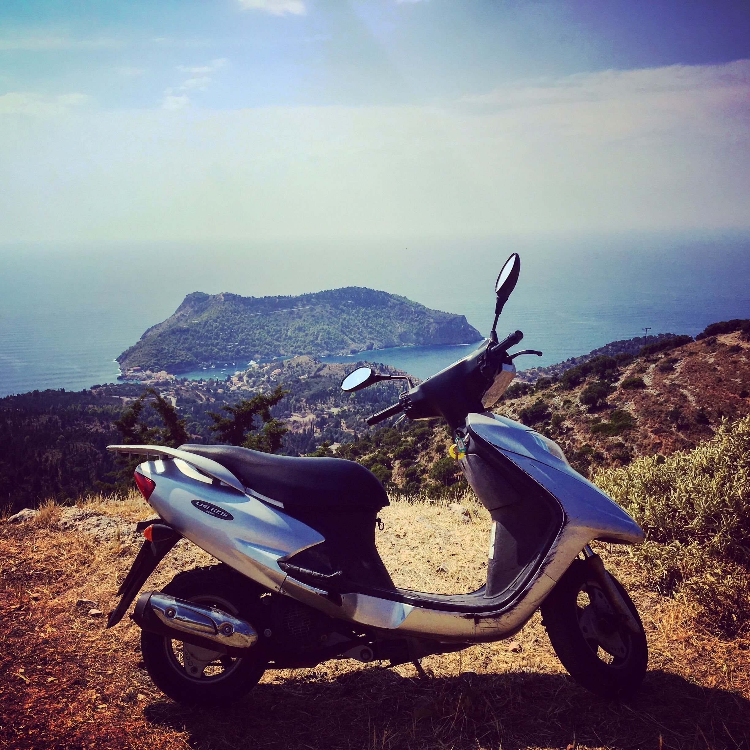 A parked scooter on a hillside overlooking a coastal town and a small island.
