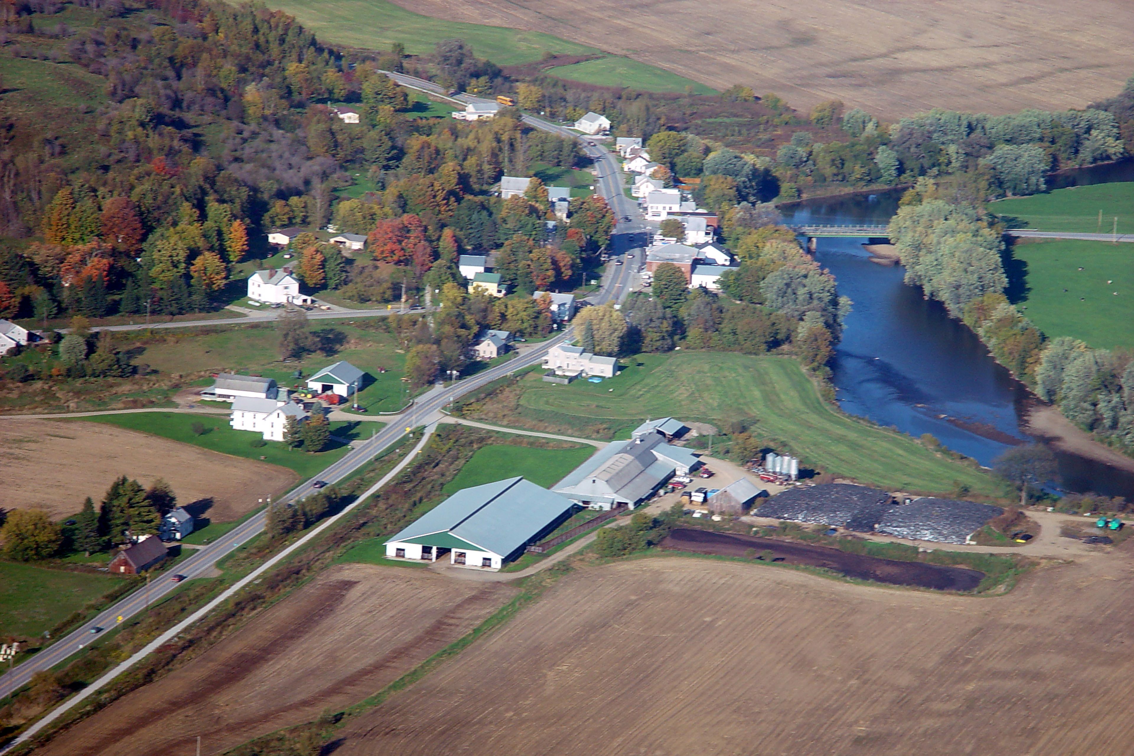 An aerial view of a small town with a river running through it, surrounded by fields and trees.