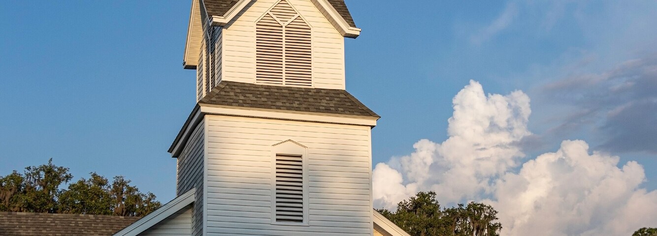 A white church with a steeple and a cross, surrounded by greenery.