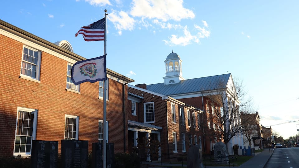 A brick building with a flagpole, a sign with a state emblem, a church steeple, and a memorial plaque.