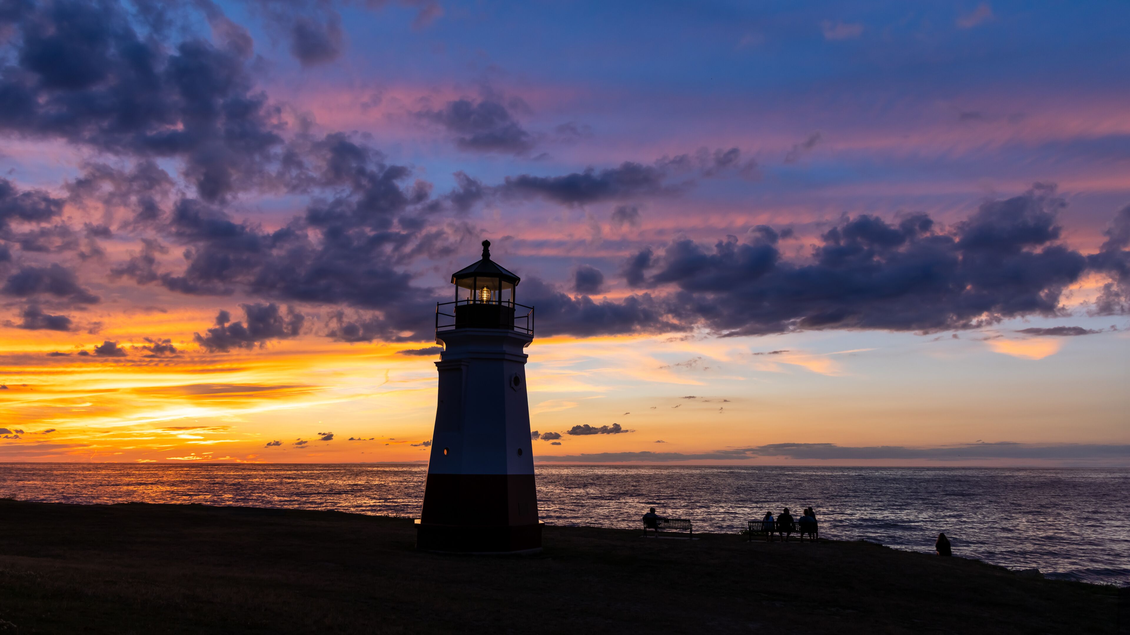 A lighthouse at sunset with a colorful sky and the ocean in the foreground.