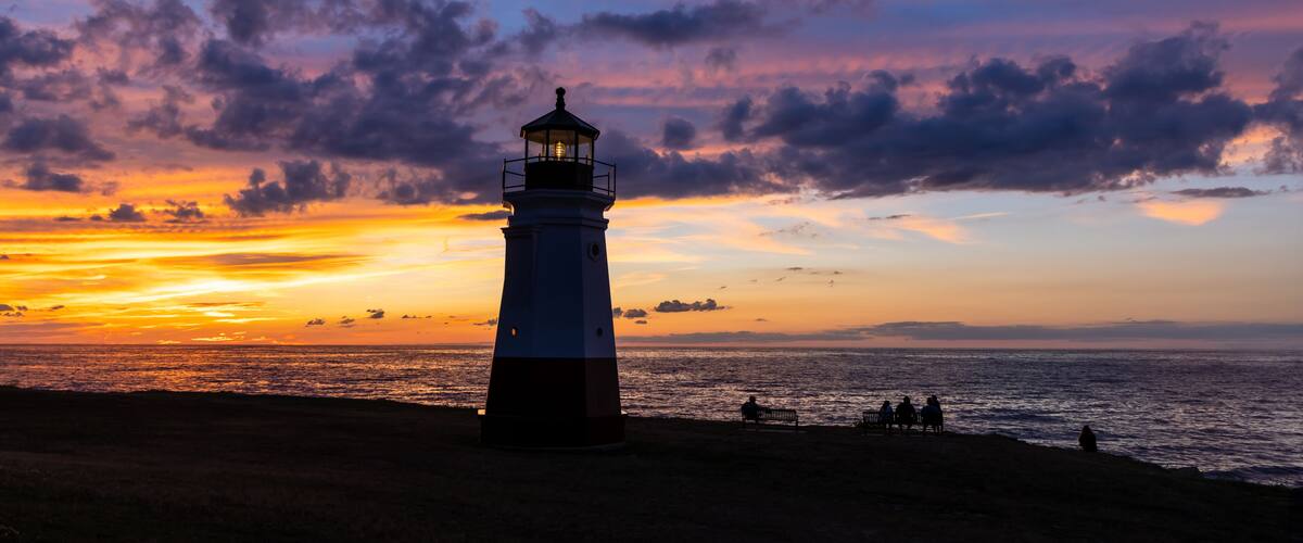 A lighthouse at sunset with a colorful sky and the ocean in the foreground.
