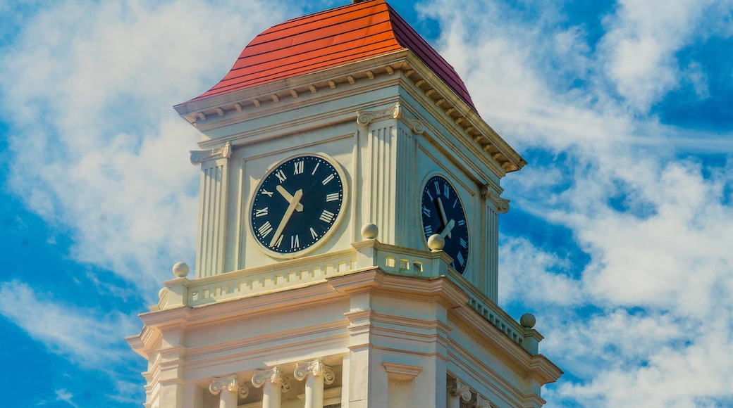 Una torre de reloj con números romanos y techo rojo, adosada a un edificio de paredes de ladrillo amarillo y columnas blancas.