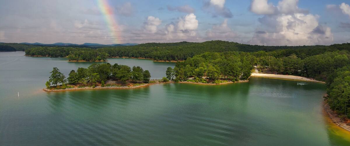 an aerial shot of a gorgeous summer landscape on the lake with rippling green water surrounded by lush green trees, grass and plants with blue sky, clouds and a rainbow at Red Top Mountain State Park