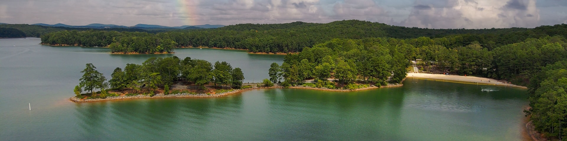 an aerial shot of a gorgeous summer landscape on the lake with rippling green water surrounded by lush green trees, grass and plants with blue sky, clouds and a rainbow at Red Top Mountain State Park