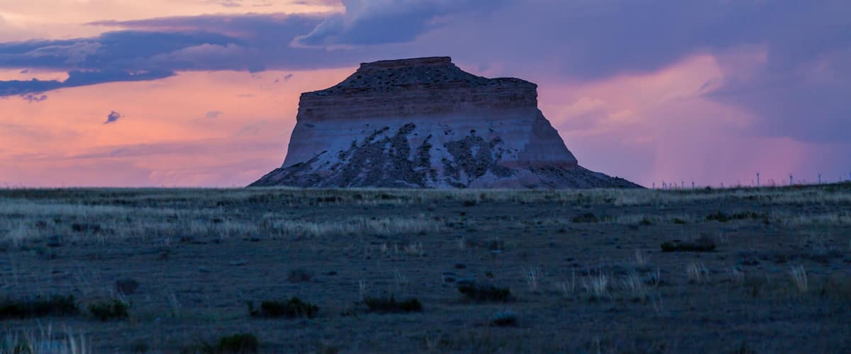 A large, flat-topped hill with sparse grass in a field, under a dramatic sky with pink and purple clouds.