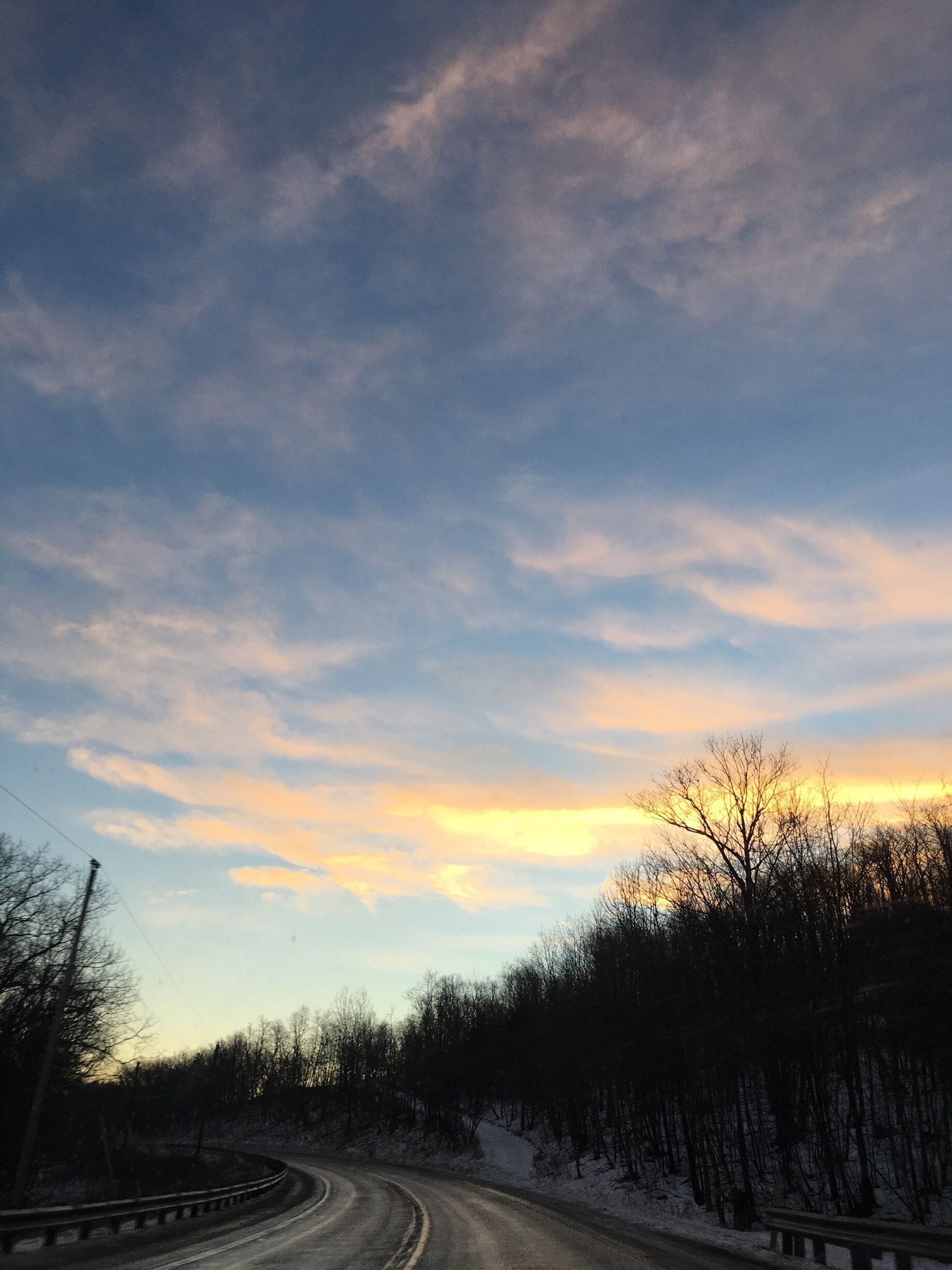 A winding road curving through a forest with bare trees and a sunset sky.