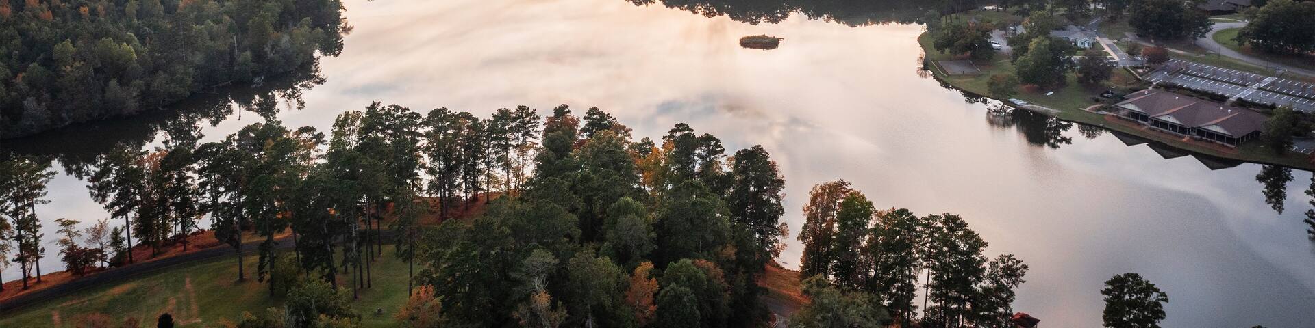 A sunset over a lake surrounded by dense forest and a distant building complex.