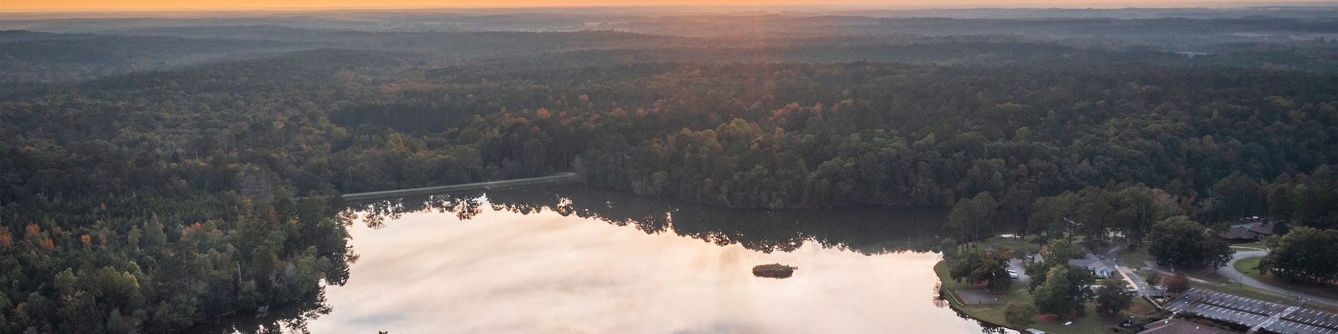 A sunset over a lake surrounded by dense forest and a distant building complex.
