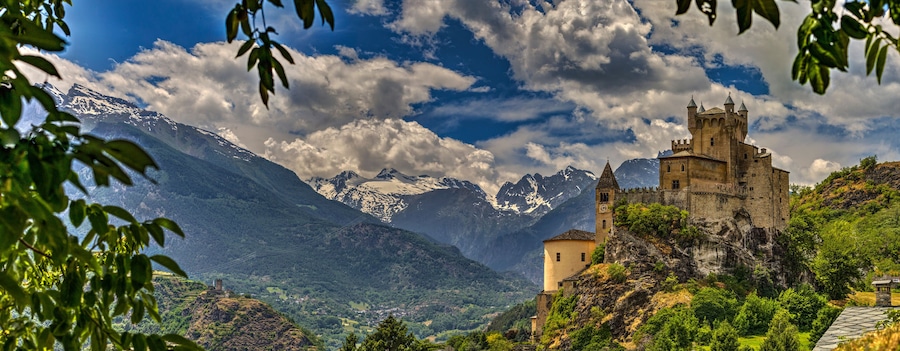 A castle on a hill with snow-capped mountains in the background.