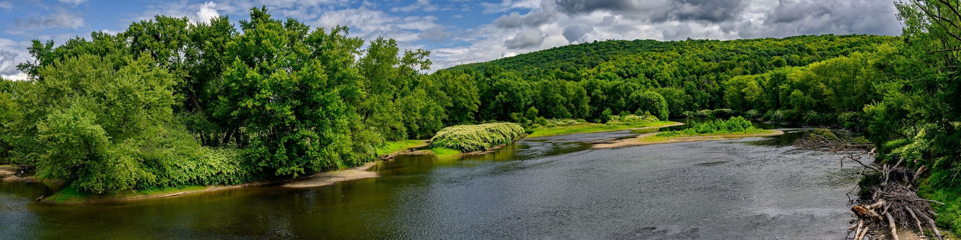 This panoramic image captures a serene river winding through a lush, green landscape under a partly cloudy sky. The far bank is covered with dense foliage, while a forested hill rises in the distance.
