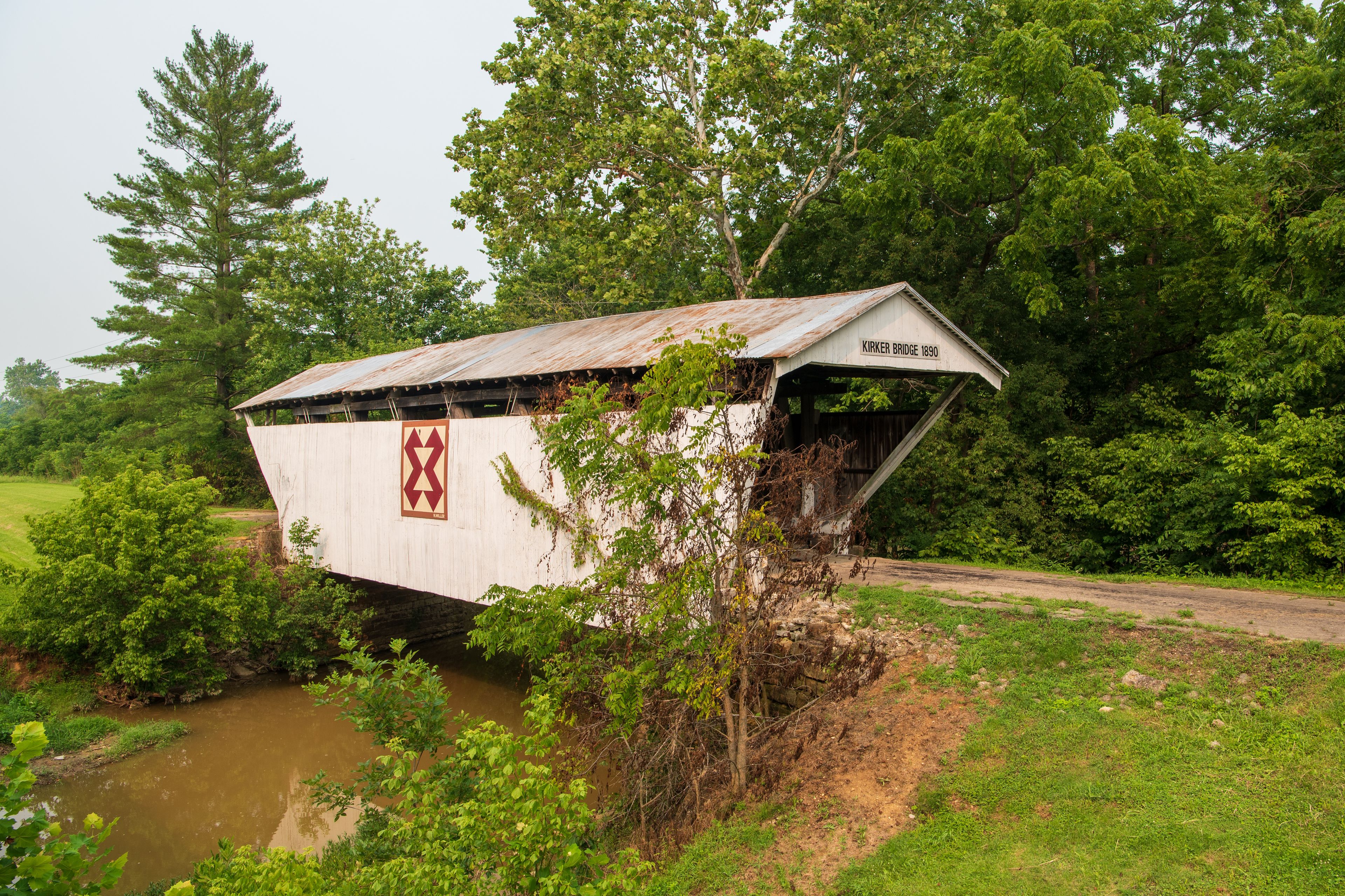 A white covered bridge with a red X on its side, labeled 'Kirkker Bridge 1890,' spanning a river in a wooded area.