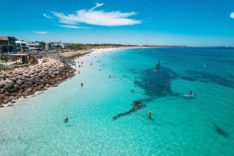 A coastal beach with clear turquoise waters, rocky shoreline, and buildings along the shore.