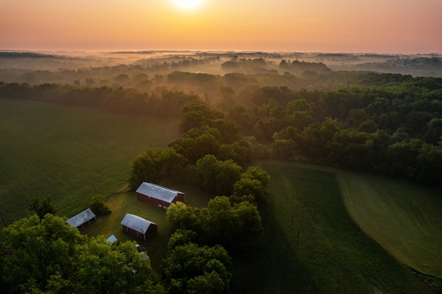 A rural landscape with a barn, trees, and fields during sunrise.