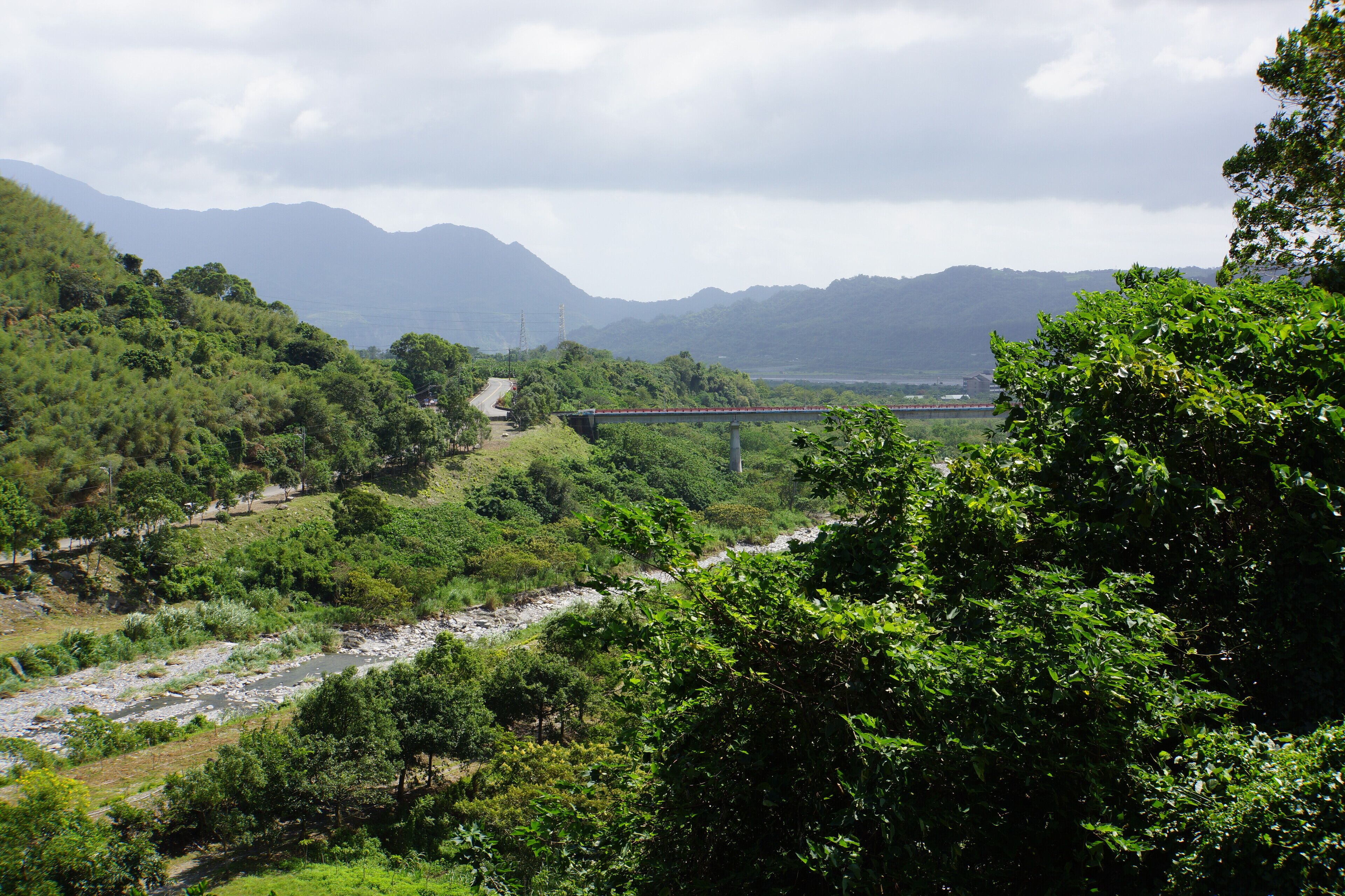 A river flowing through a lush valley with a bridge in the distance and mountains in the background.