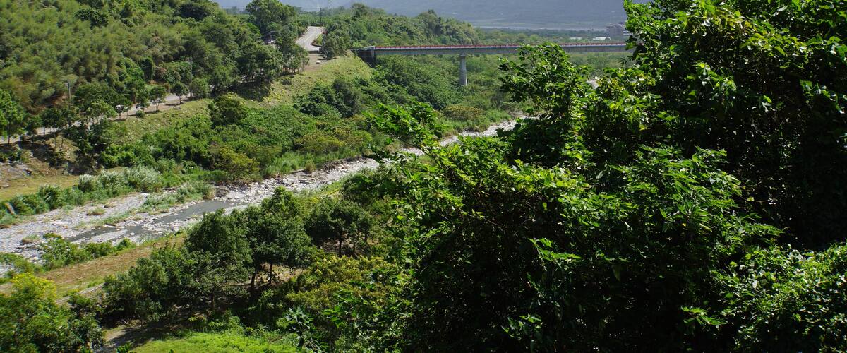 A river flowing through a lush valley with a bridge in the distance and mountains in the background.