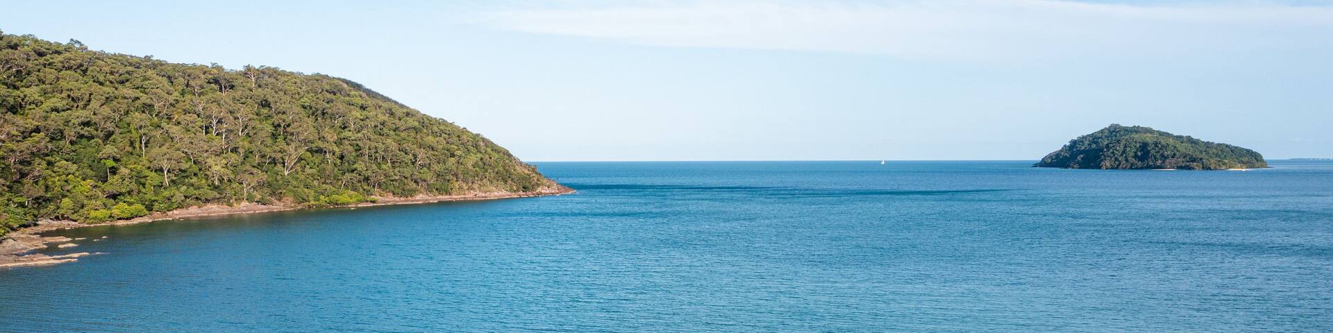 Un paisaje costero con una playa de arena, aguas azules tranquilas y una colina boscosa al fondo.