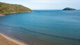Eine Küstenlandschaft mit sandigem Strand, ruhigem blauen Wasser und einem bewaldeten Hügel im Hintergrund.