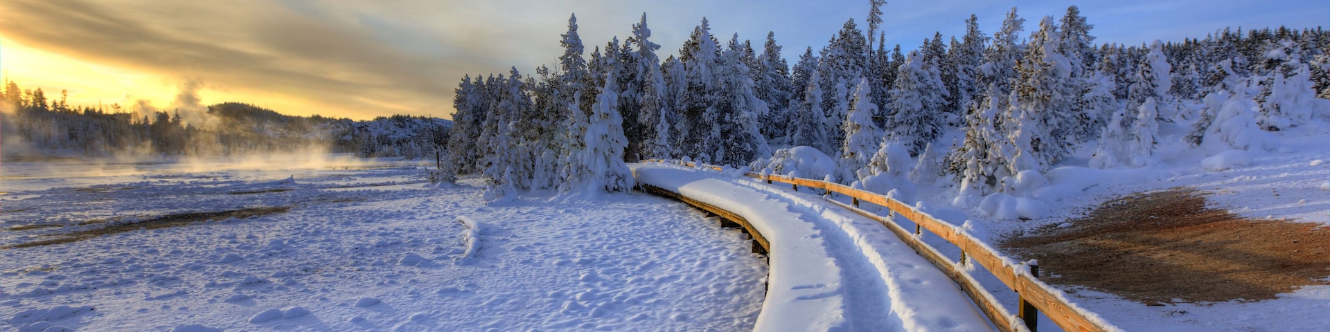 Schneebedeckte Landschaft mit einem Holzzaun, der sich einen Weg entlang schlängelt, dichten Nadelbäumen und einem sich spiegelnden Gewässer.