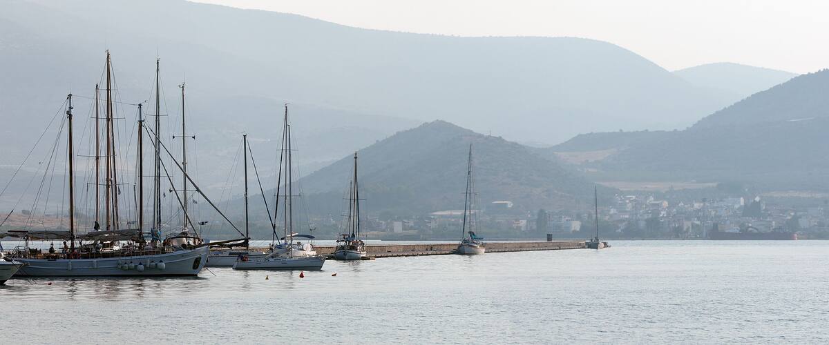 A harbor with sailboats docked, a pier, and a town in the background with mountains.