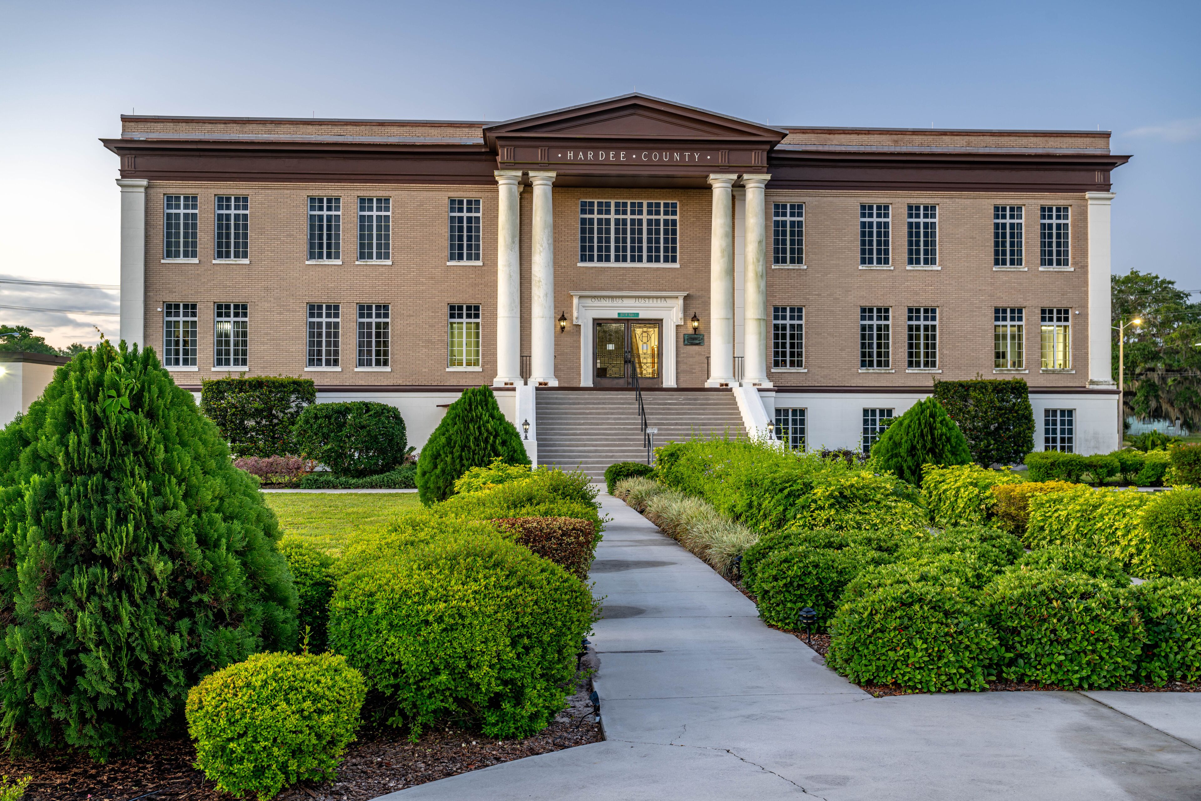A brick courthouse with columns, a central entrance, and a well-maintained garden with various shrubs.