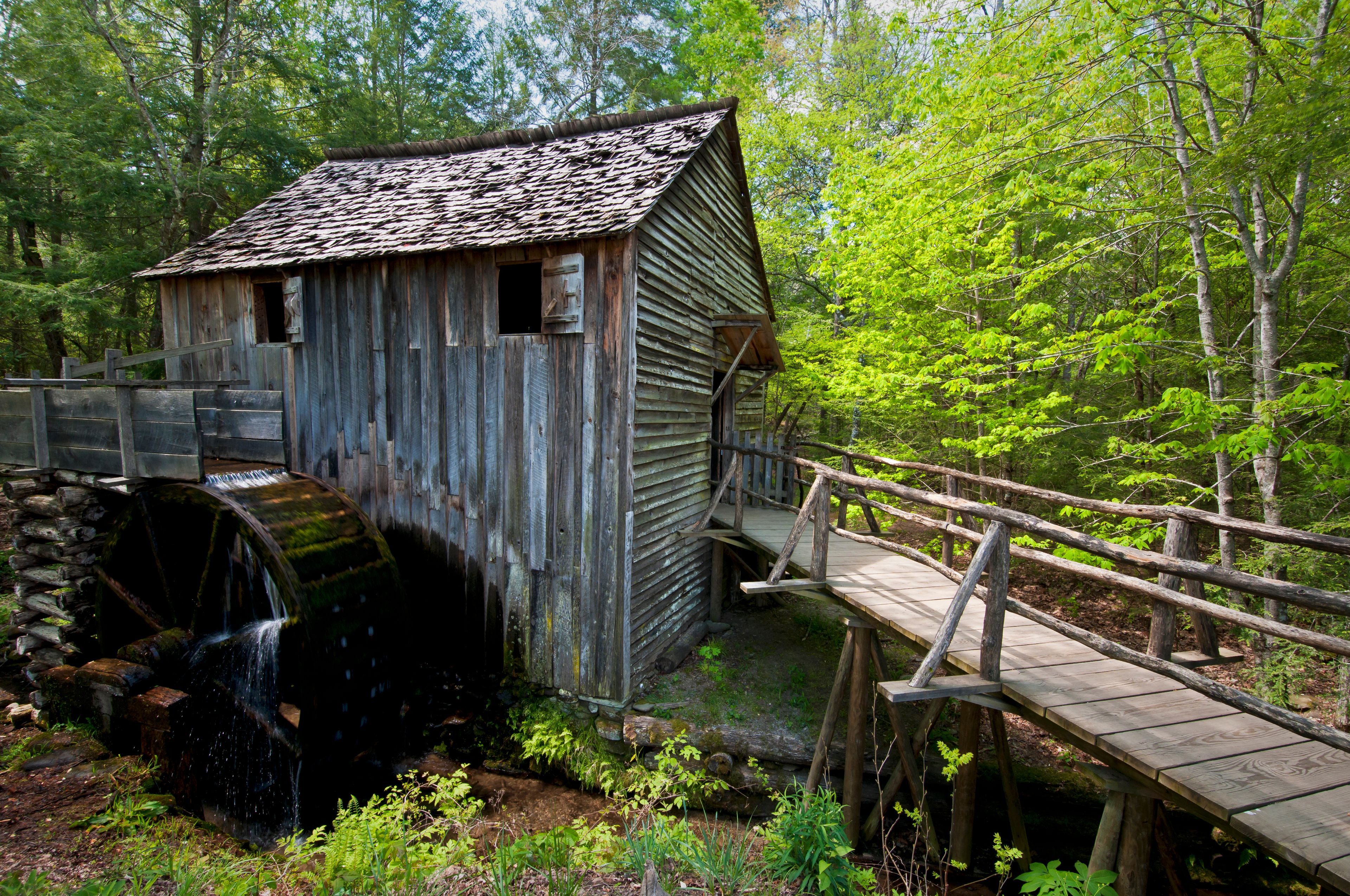 A rustic wooden mill with a water wheel, surrounded by lush greenery and a wooden bridge.