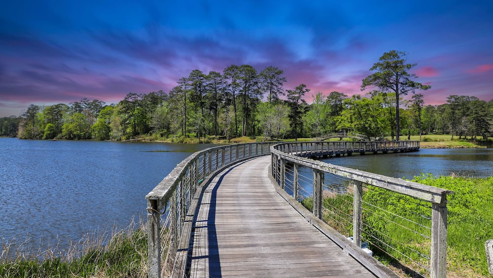 A long winding brown wooden bridge over a rippling blue lake surrounded by lush green trees, grass and plants with blue sky and powerful clouds at sunset at Callaway Gardens in Pine Mountain Georgia