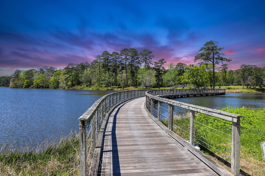 A long winding brown wooden bridge over a rippling blue lake surrounded by lush green trees, grass and plants with blue sky and powerful clouds at sunset at Callaway Gardens in Pine Mountain Georgia