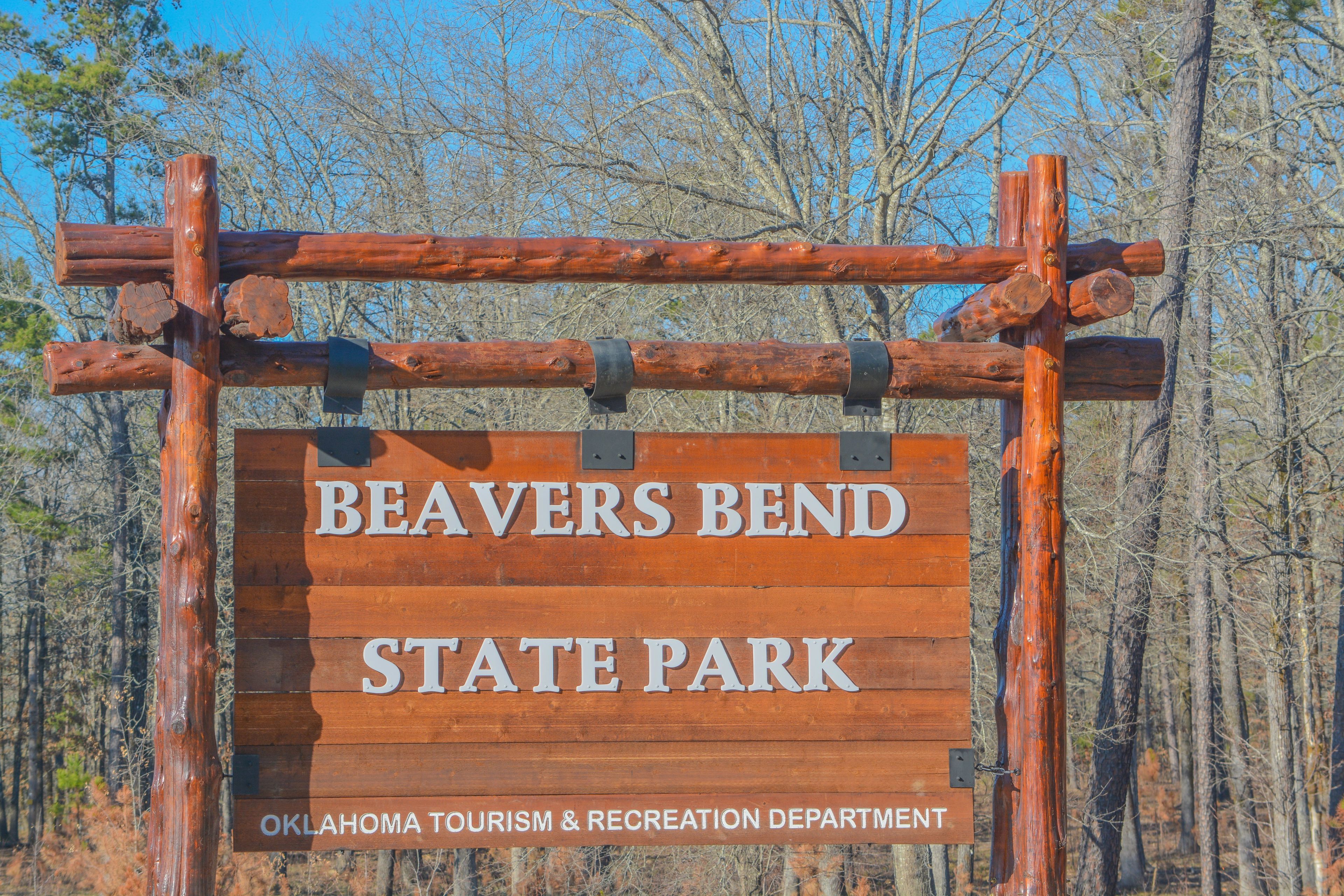 A wooden sign with the text 'Beavers Bend State Park' and 'Oklahoma Tourism & Recreation Department' against a backdrop of trees.
