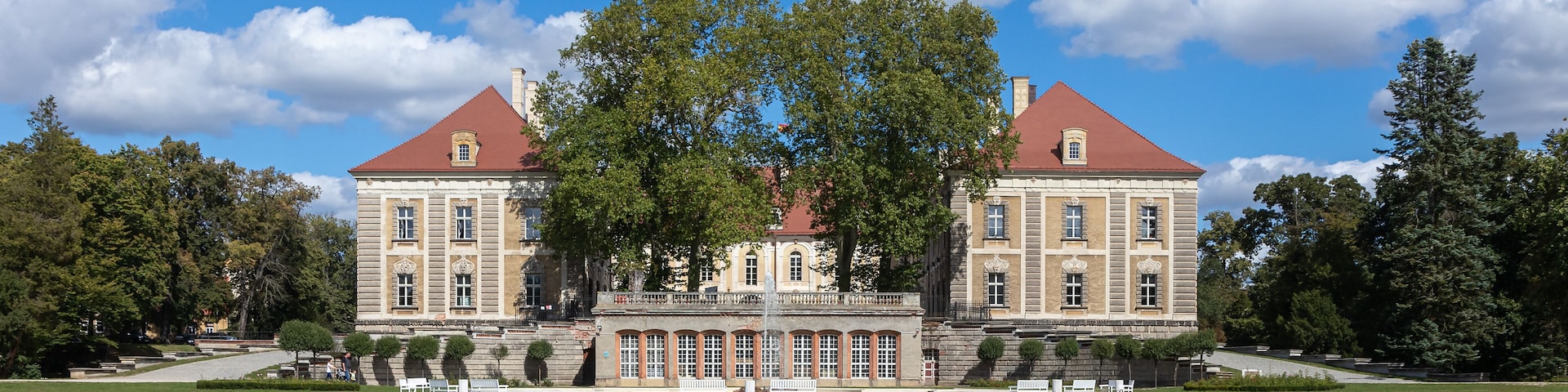 Ein großes, historisches Gebäude mit rotem Dach und symmetrischer Gestaltung, umgeben von einem gepflegten Rasen und Bäumen.