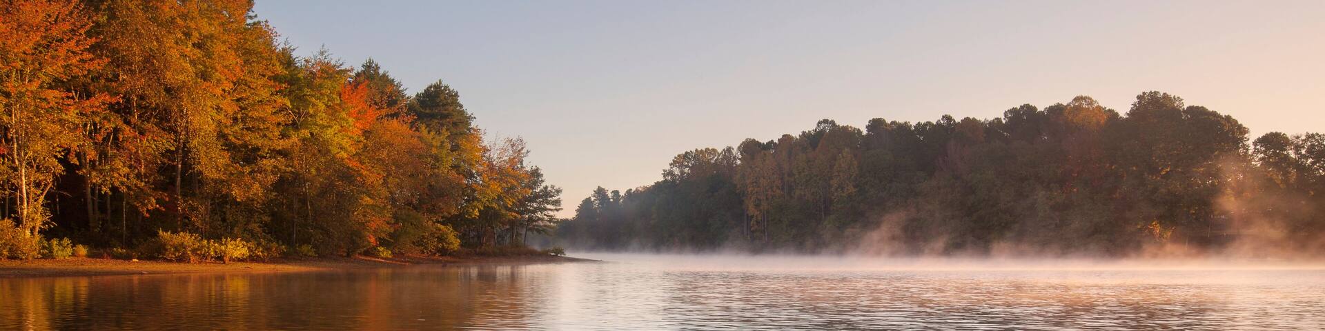 A calm lake with autumn foliage, mist over the water, and a rocky shoreline.