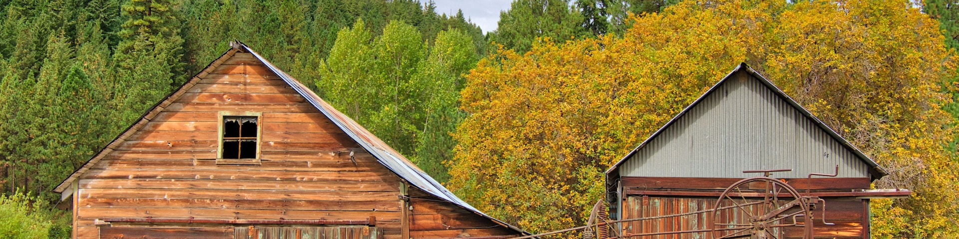 A rustic wooden barn with a large wheel mechanism, surrounded by a forest with autumn-colored trees.