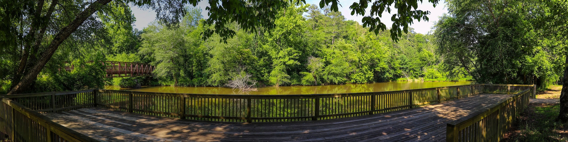 A wooden boardwalk curving around a pond, surrounded by dense green foliage and a metal bridge in the background.