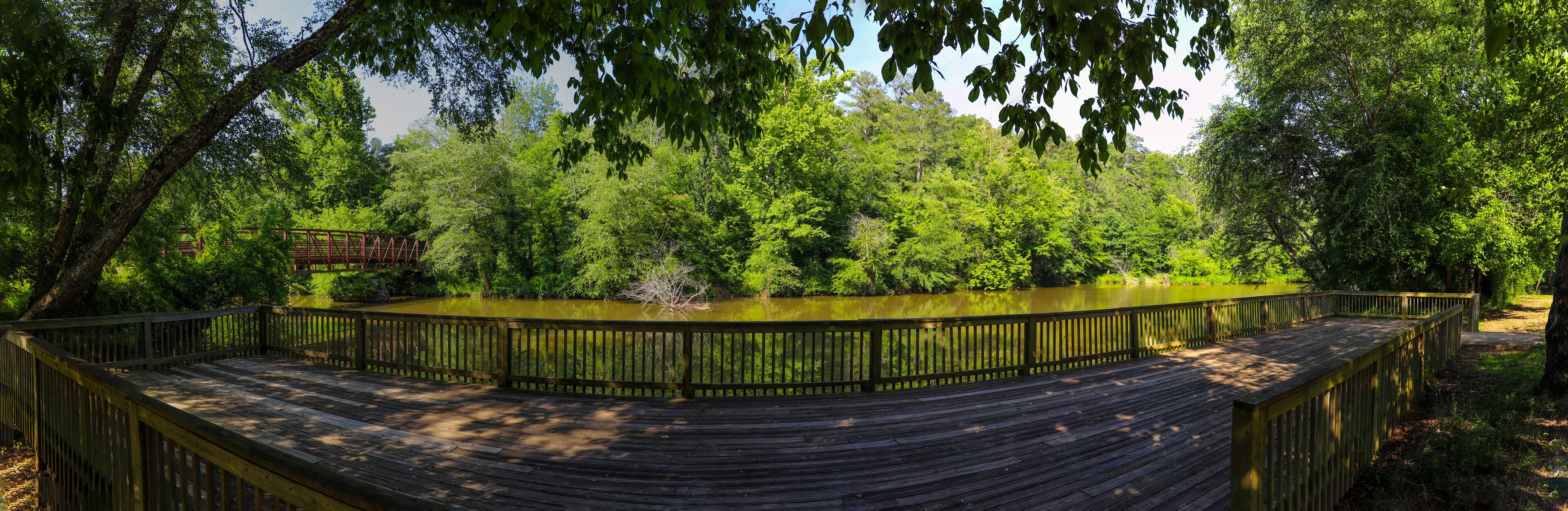 A wooden boardwalk curving around a pond, surrounded by dense green foliage and a metal bridge in the background.