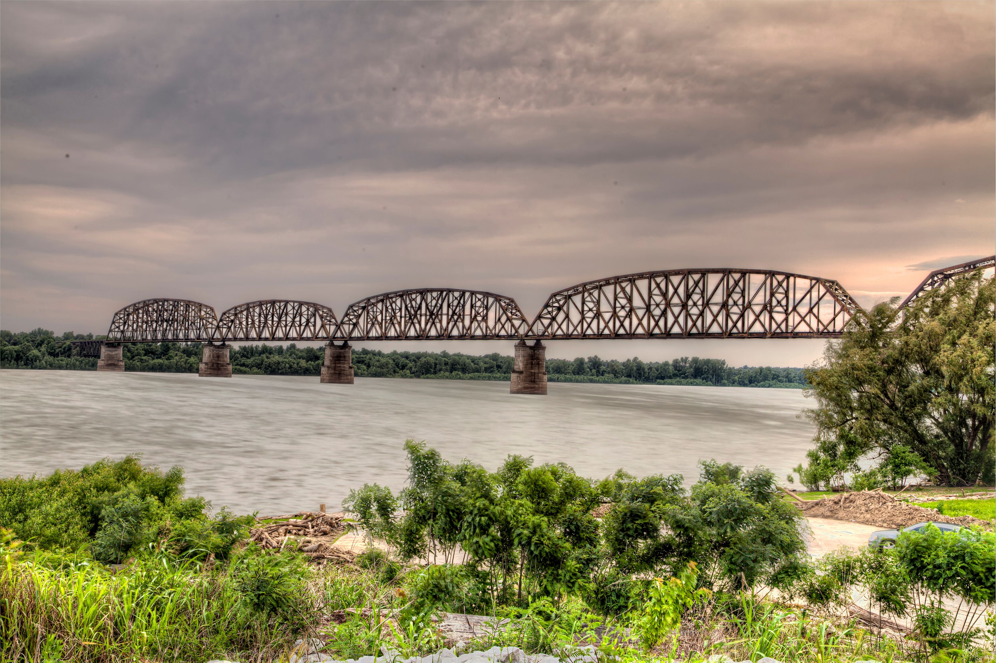 A steel bridge over a river with a cloudy sky.
