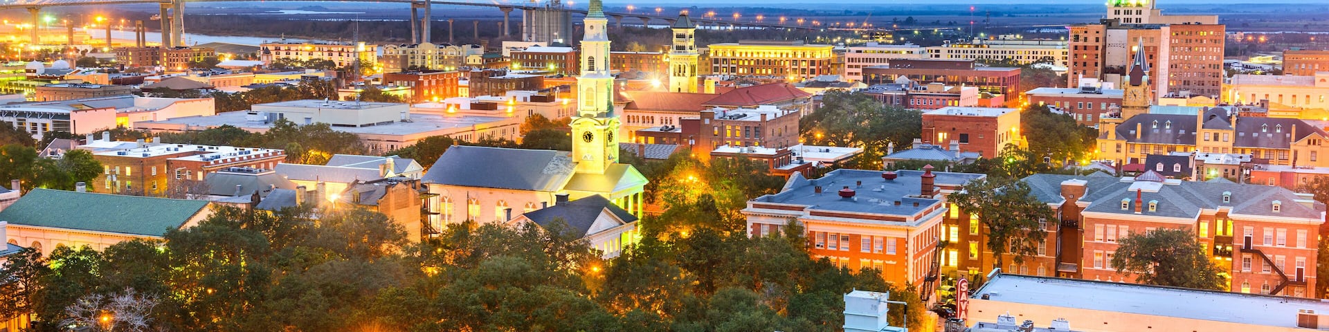 A cityscape at dusk with illuminated buildings, a prominent church spire, and a bridge in the background.