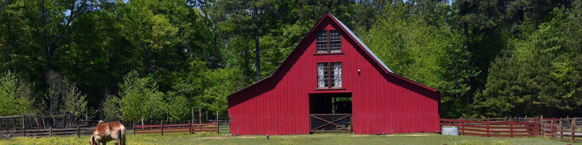 Ein rotes Bauernhaus, umgeben von einem Holzzaun, mit einem weidenden Pferd im Vordergrund.