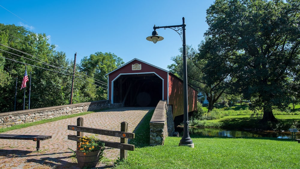A covered bridge with a sign, a wooden bench, a flower pot, a lamp post, and a pond.