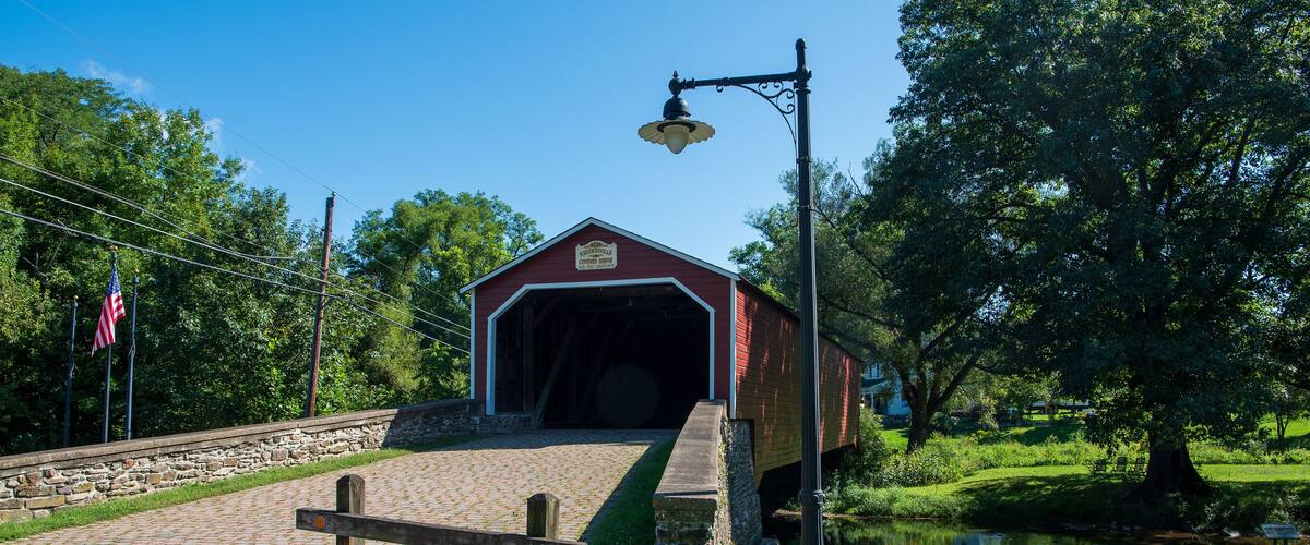 A covered bridge with a sign, a wooden bench, a flower pot, a lamp post, and a pond.