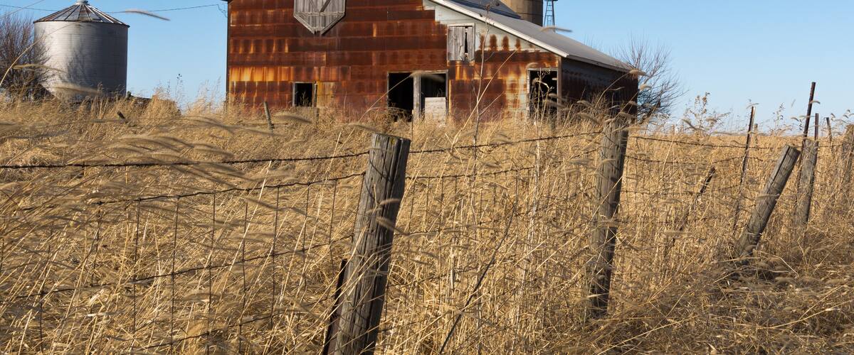 A rusted barn with a silo and a fence in a dry field.