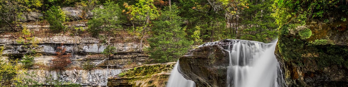A waterfall cascading over a rocky cliff into a calm pool surrounded by forested terrain.