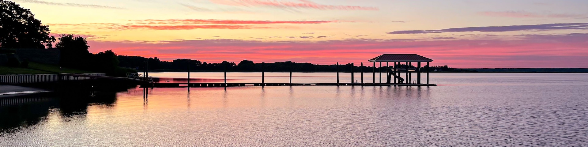 A sunset over a calm lake with a pier and a gazebo.