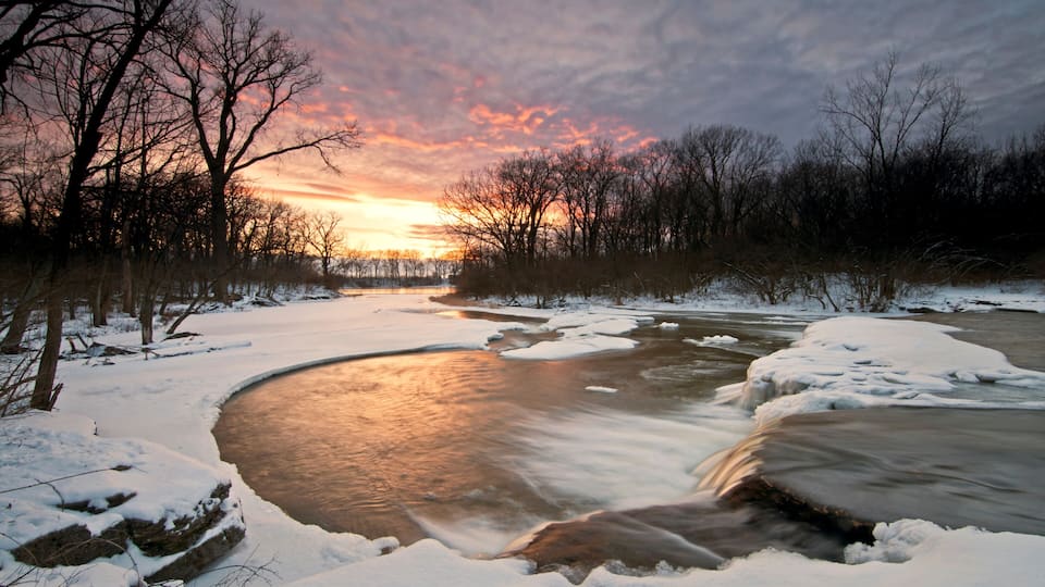 A frozen river with snow-covered banks and trees in the background during sunset.