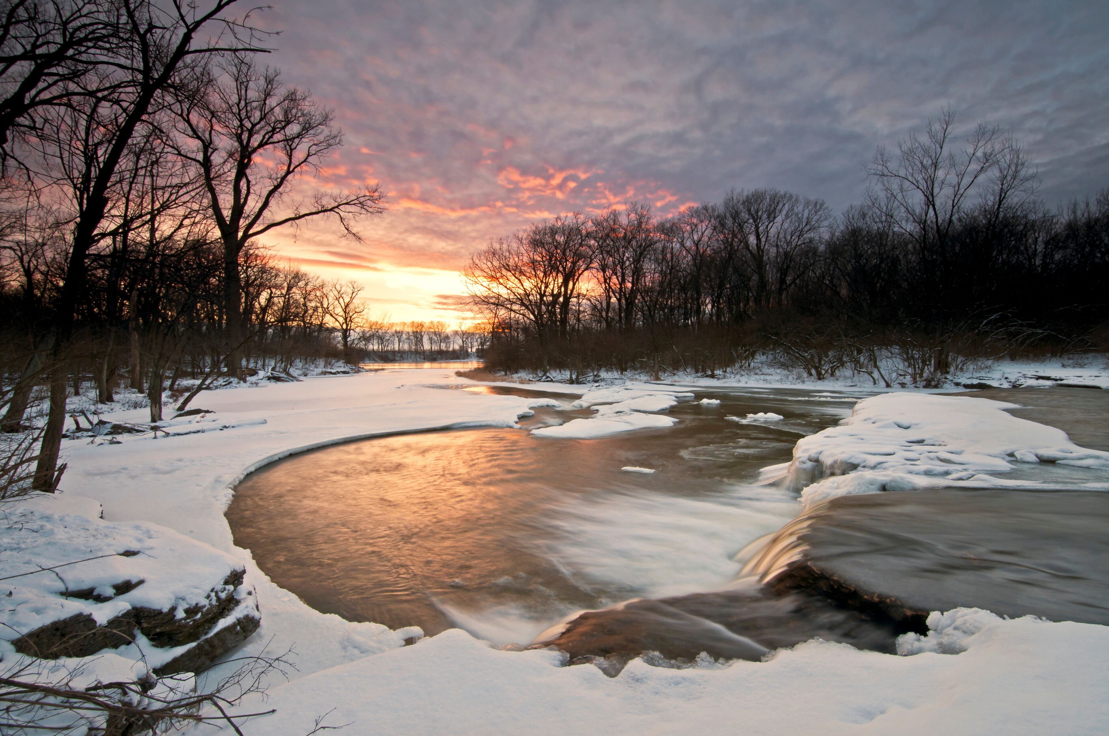 A frozen river with snow-covered banks and trees in the background during sunset.
