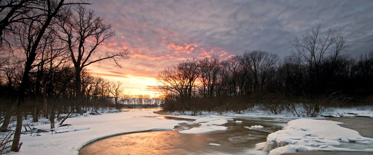 A frozen river with snow-covered banks and trees in the background during sunset.
