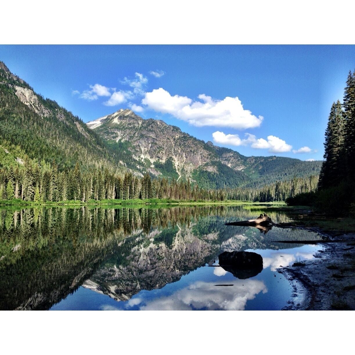Um lago de montanha com reflexos nítidos, rodeado por florestas densas e montanhas rochosas.